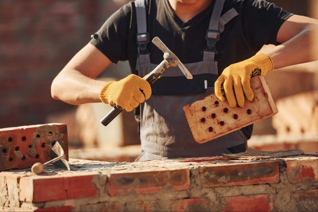 Cropped shot of a skilled mason in overalls preparing bricks with a hammer.